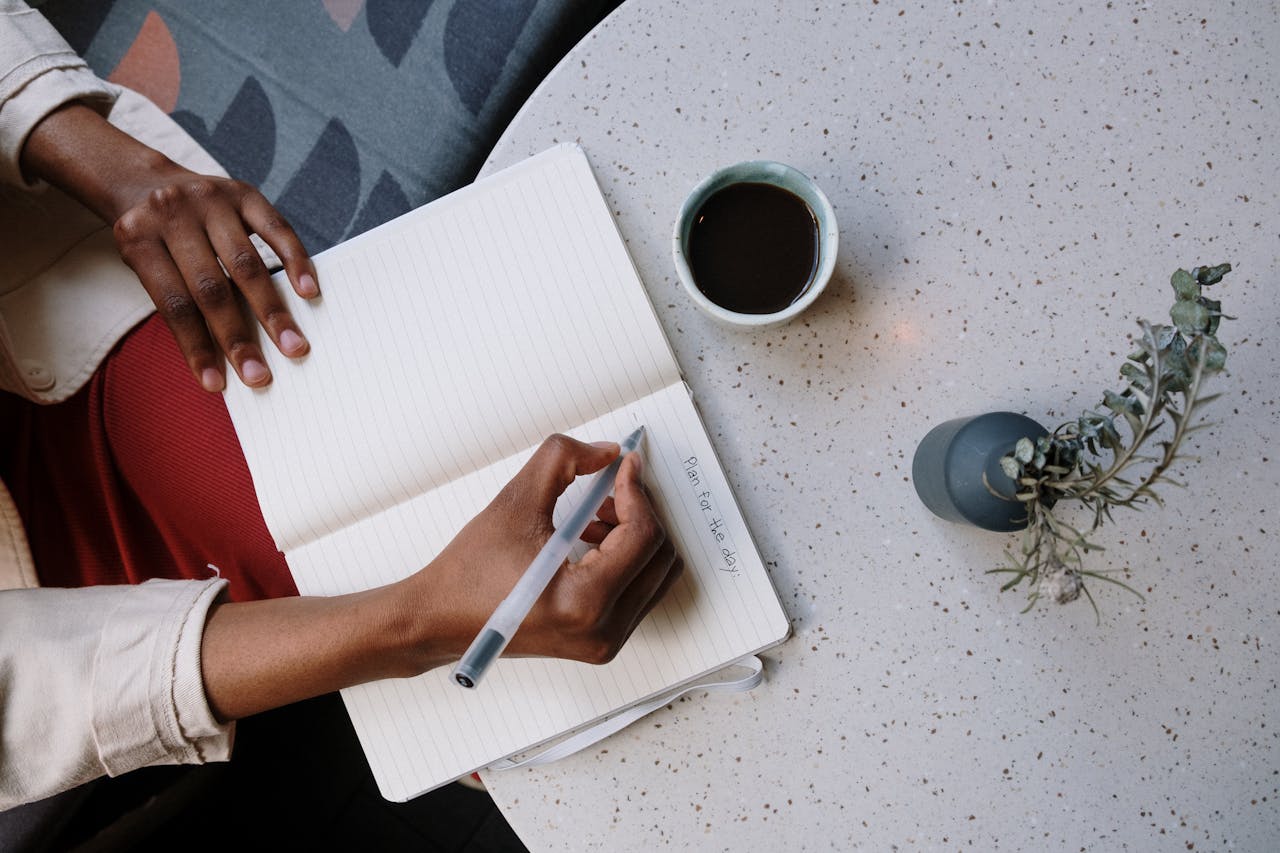 our-services-2 Person writing in a planner with coffee at a café, focusing on planning and productivity.